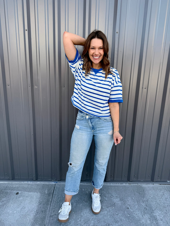 Woman wearing a blue and white striped shirt and light blue jeans standing against a gray wall.