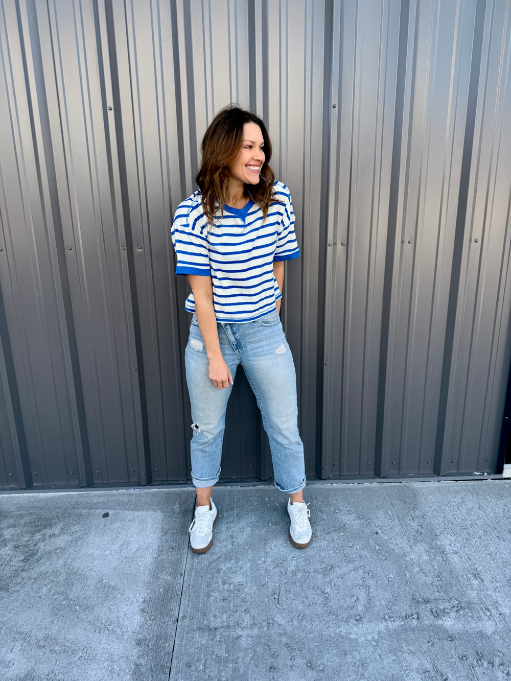 Woman wearing a striped shirt and jeans standing against a gray wall.