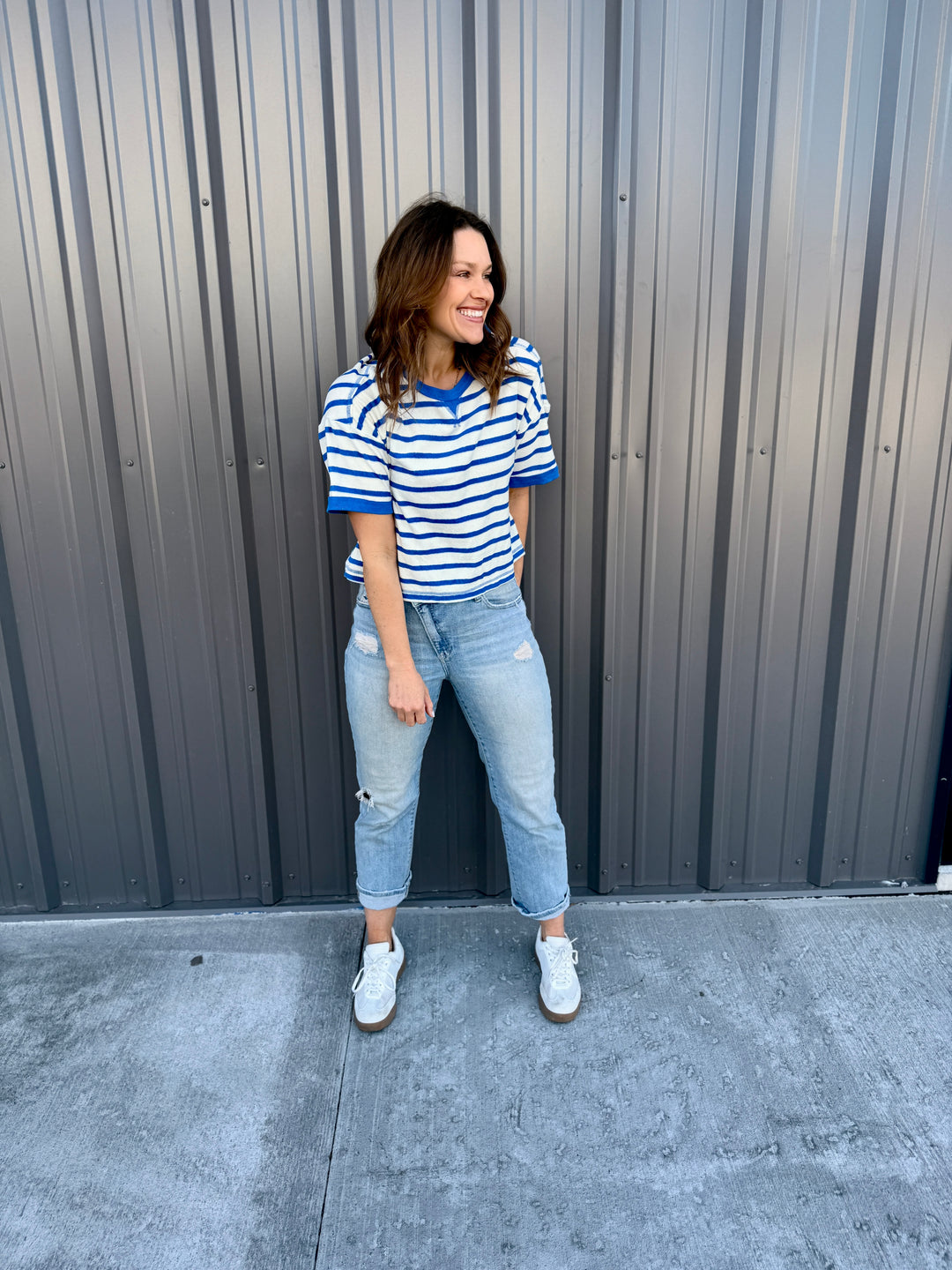 Woman wearing a striped shirt and jeans standing against a gray wall.