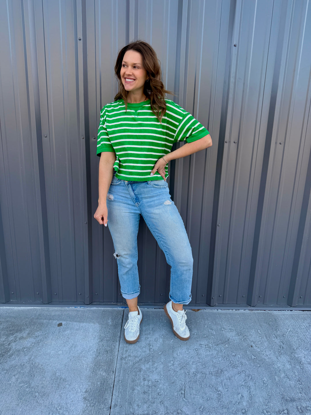 Woman wearing a green and white striped shirt and light blue jeans standing against a gray wall.