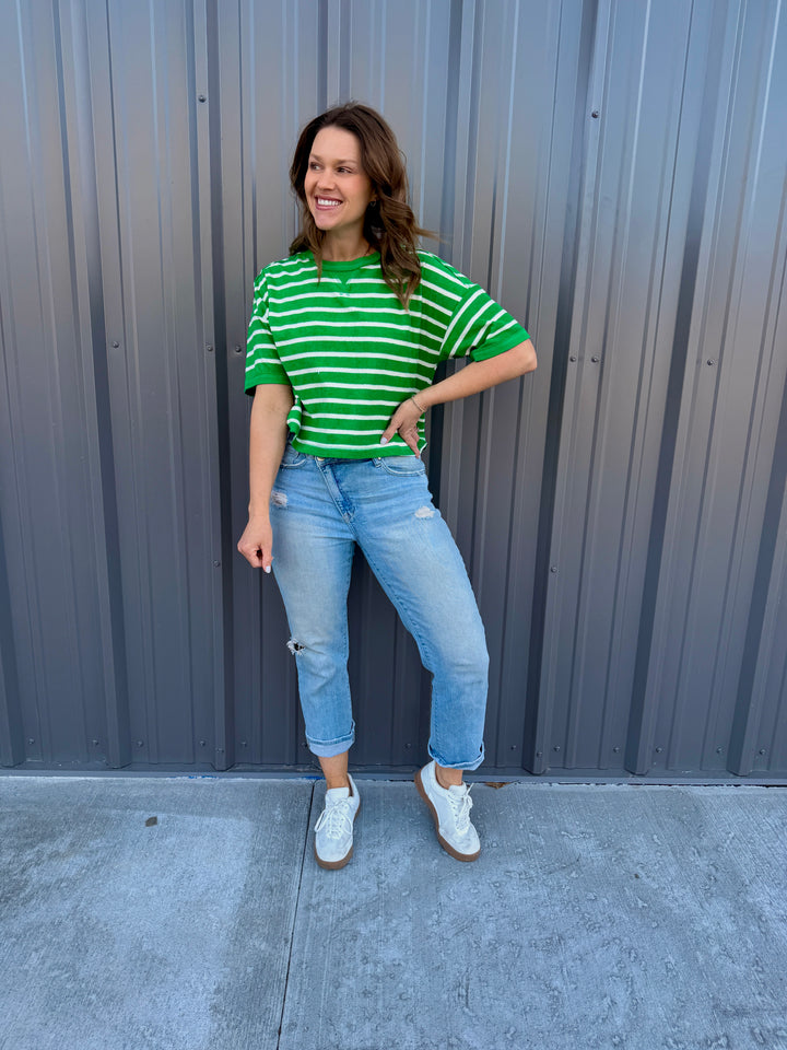 Woman wearing a green and white striped shirt and light blue jeans standing against a gray wall.